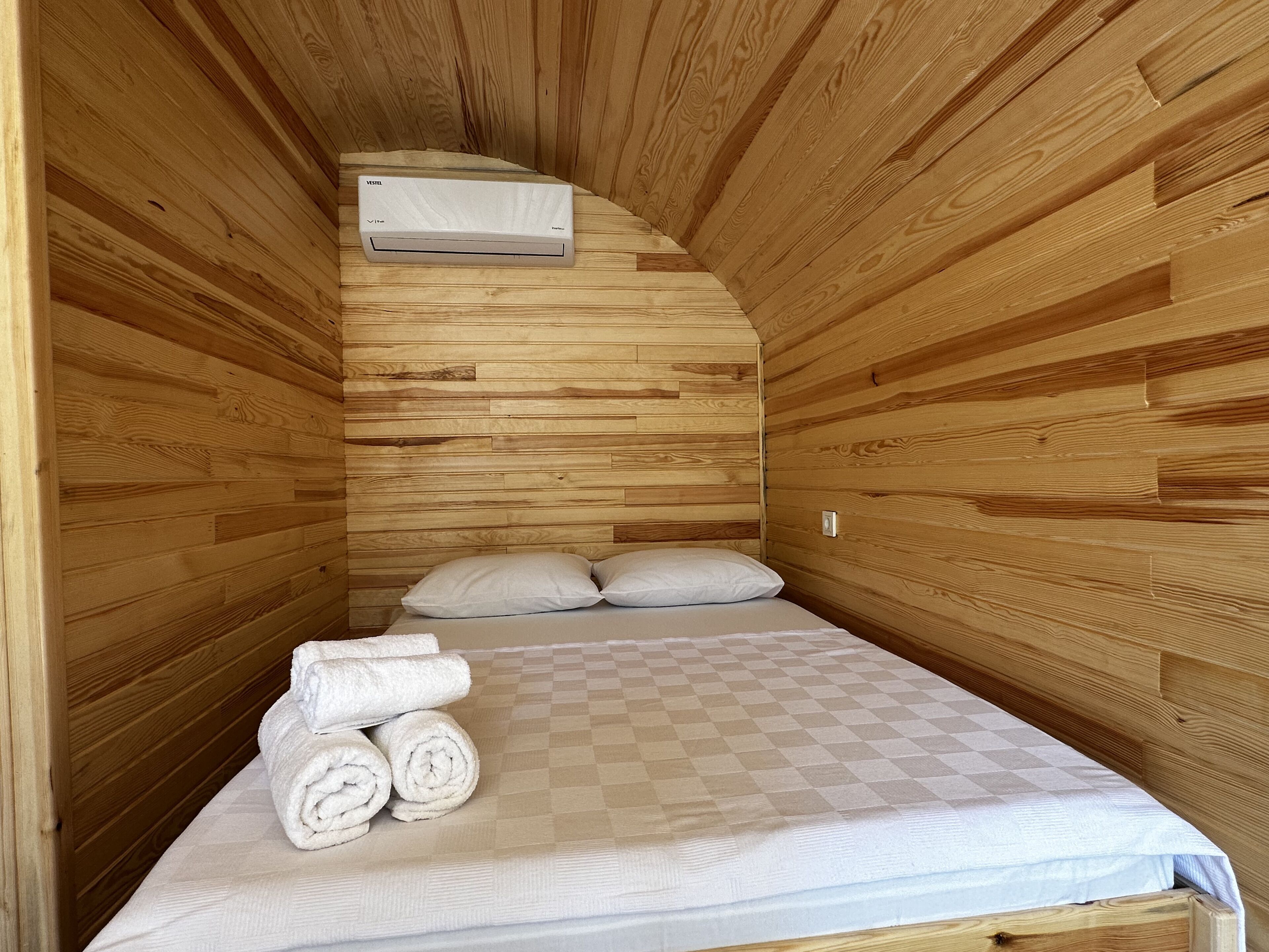 A small, cozy room with wooden walls, a single bed with a checkered bedspread, two pillows, and a stack of rolled towels.