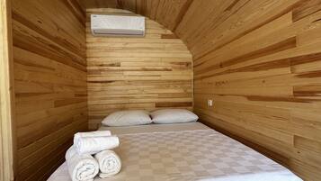 A small, cozy room with wooden walls, a single bed with a checkered bedspread, two pillows, and a stack of rolled towels.