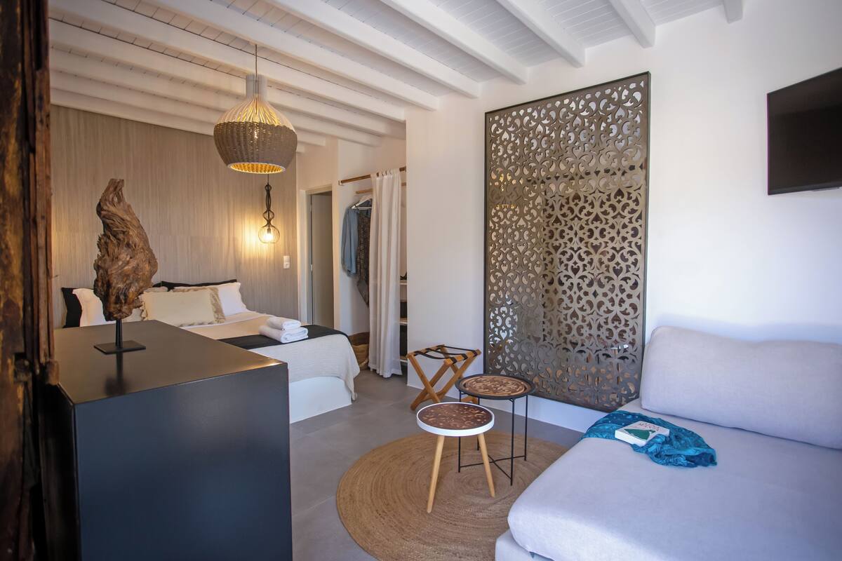 A modern hotel room with a bed, a sofa, two chairs, a decorative wall panel, and a hanging light fixture.