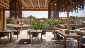 An outdoor dining area with stone flooring, wooden tables, and chairs, overlooking a pool and stone buildings. - Nur Mykonian Suites (Mykonos)
