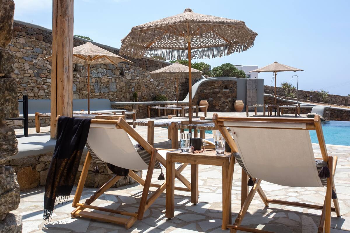 A poolside area with wooden lounge chairs, straw umbrellas, and a stone wall backdrop.