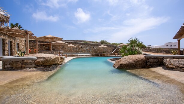 A clear pool with stone walls and umbrellas, surrounded by stone buildings and palm trees. - Nur Mykonian Suites (Mykonos)