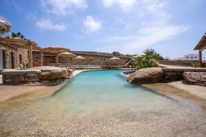 A clear pool with stone walls and umbrellas, surrounded by stone buildings and palm trees. - Nur Mykonian Suites (Mykonos)