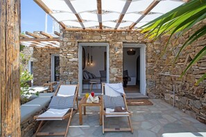 A stone-walled outdoor seating area with lounge chairs and a table, connected to a covered patio with a wooden pergola. - Nur Mykonian Suites (Mykonos)