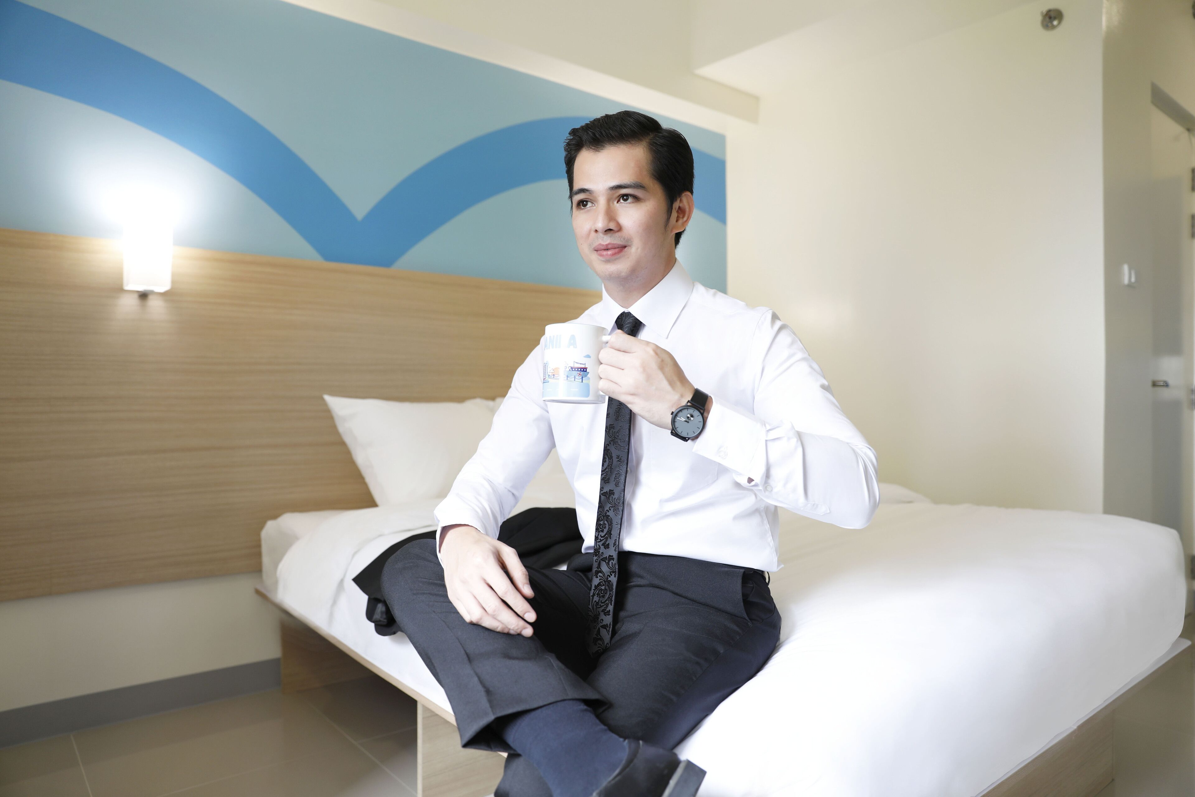 A man sitting on a bed in a hotel room with a blue and white wall design, a wooden headboard, and a white pillow.