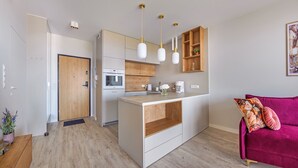 A modern kitchen with wooden cabinets, a central island, pendant lights, and a red sofa in the living area. - Apartamenty Sun&Snow Modern Tower (Gdynia)