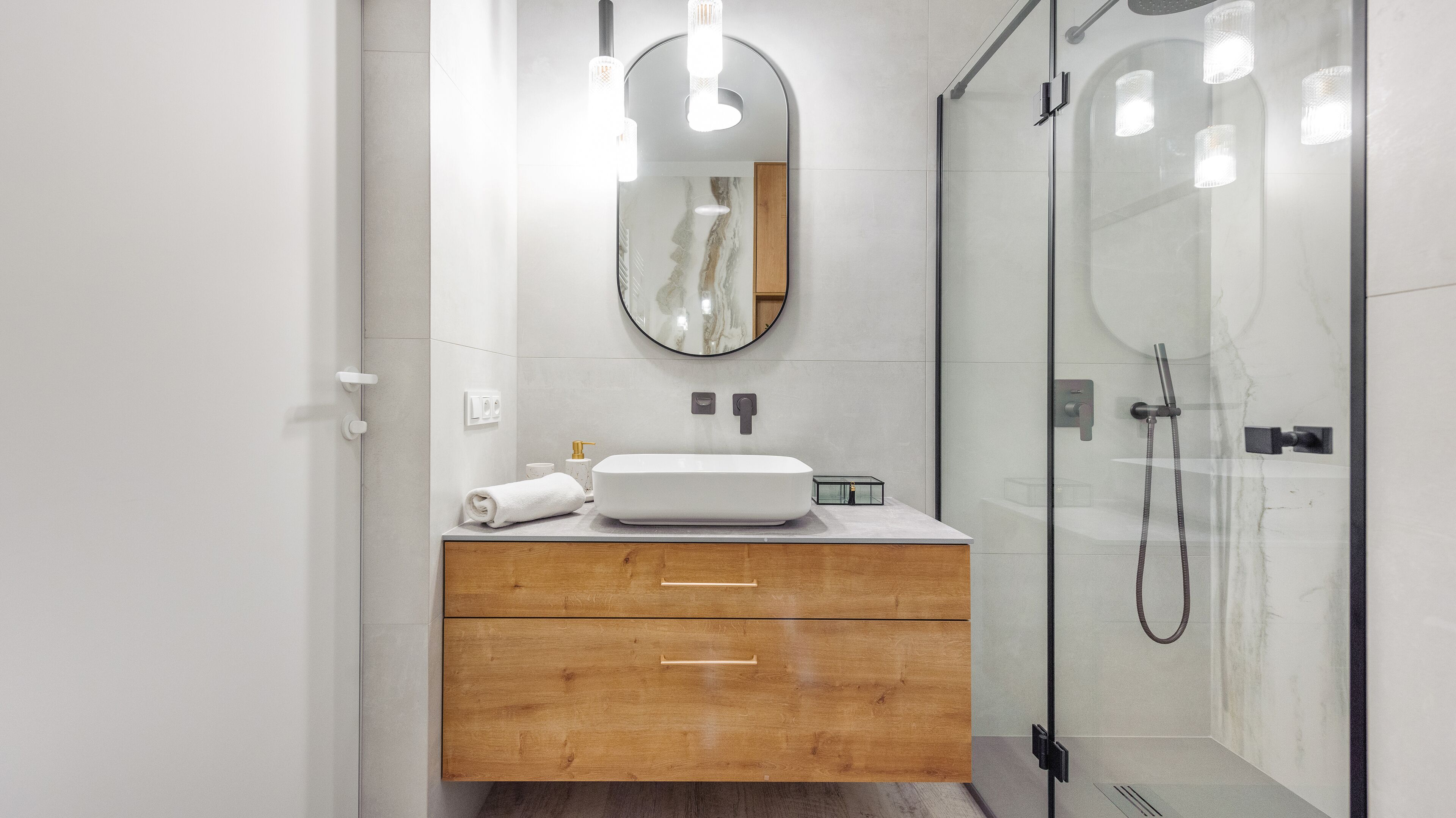 A modern bathroom with a glass shower enclosure, a wooden vanity with a white sink, and a round mirror.