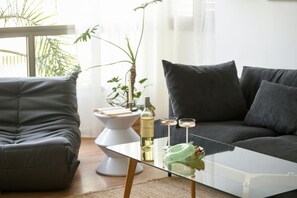 A modern living room with a glass coffee table, two grey sofas, a white side table, and a bottle of wine. - Stylish 2-Bedroom Green Oasis (Tel Aviv)
