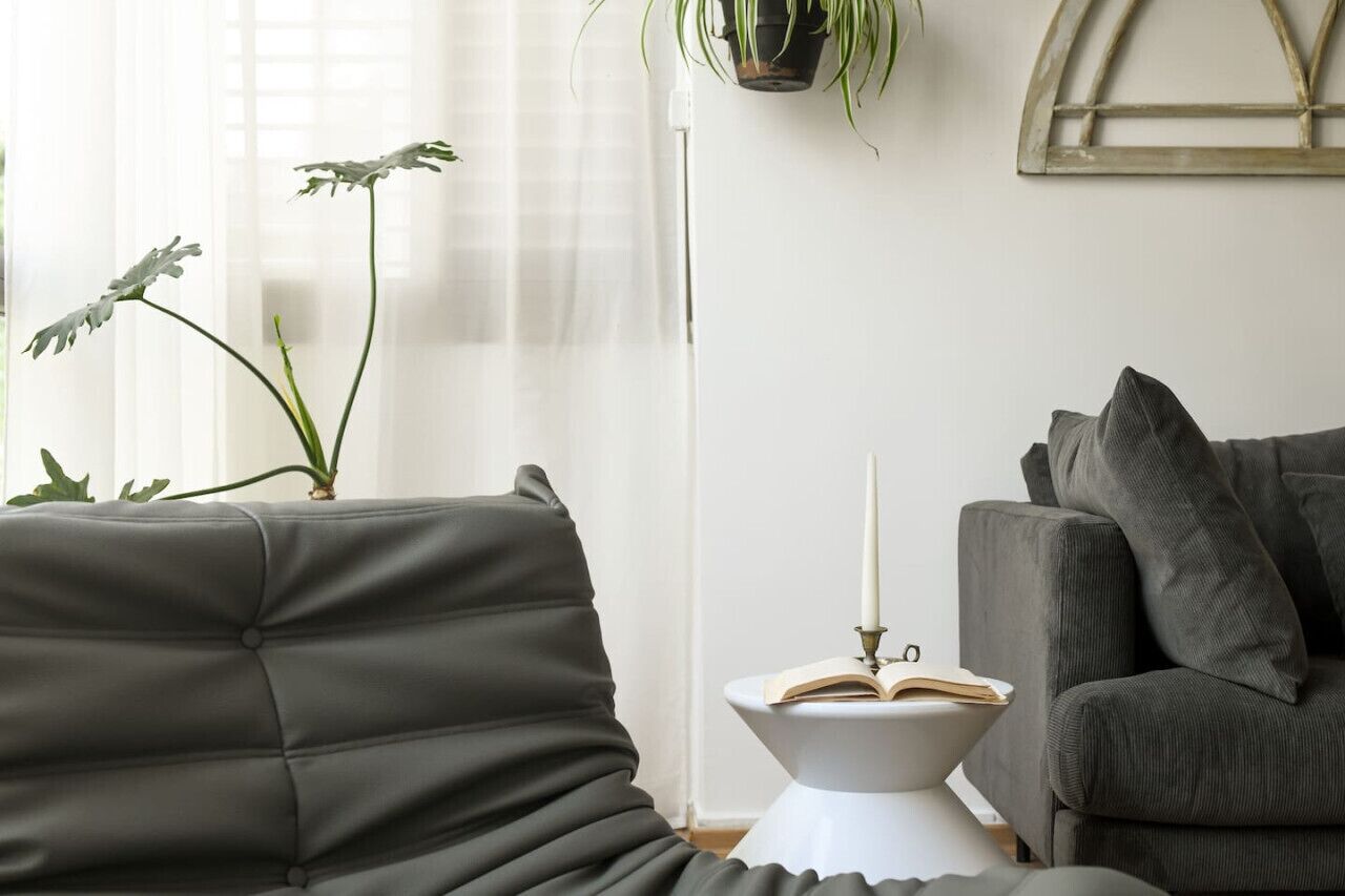 Un salon moderne avec un canapé gris, une table d’appoint blanche sur laquelle se trouvent un livre et une bougie, et une plante en pot.