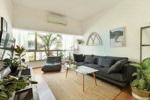 A living room with a grey sofa, a glass coffee table, and a large window with plants. - Stylish 2-Bedroom Green Oasis (Tel Aviv)