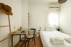 A hotel room with a bed, a desk with a chair, a small table with a lamp, a hat on a rack, and an air conditioning unit. - Stylish 2-Bedroom Green Oasis (Tel Aviv)
