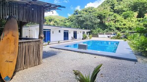 A pool area with a wooden deck, a surfboard, and a building with blue doors. - VIVE EL VALLE Hotel (Samaná)