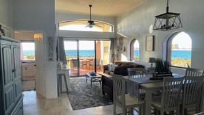 A spacious living area with a view of the sea, a dining area with white chairs, and a ceiling fan.