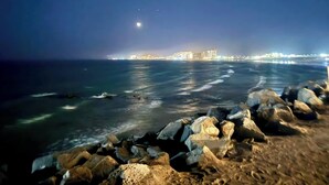 A nighttime coastal scene with a rocky foreground, calm sea, and a distant illuminated cityscape.