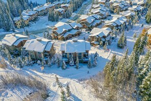 Une station de ski recouverte de neige, avec plusieurs bâtiments de style chalet disposés en cercle, entourée d’arbres à feuilles persistantes.