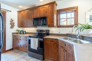 A kitchen with wooden cabinets, a black microwave, a stainless steel sink, and a window with a view of trees. - Location, Location, Location! Ski in Ski out at Trapper's Landing #23 (Sun Peaks)