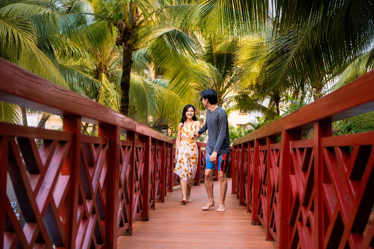 A wooden bridge with red railings, surrounded by palm trees.