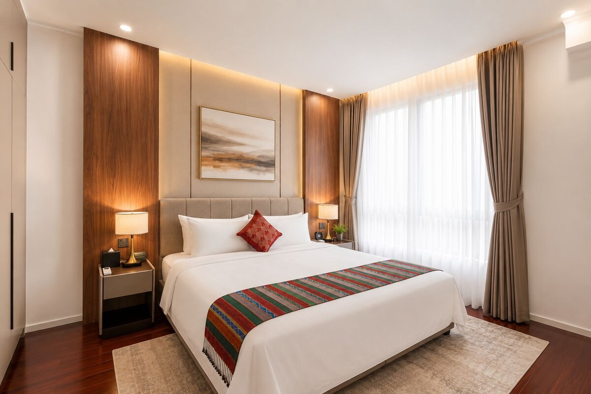 A modern hotel room with a large bed, bedside lamps, a painting on the wall, and a view of the outdoors through sheer curtains.