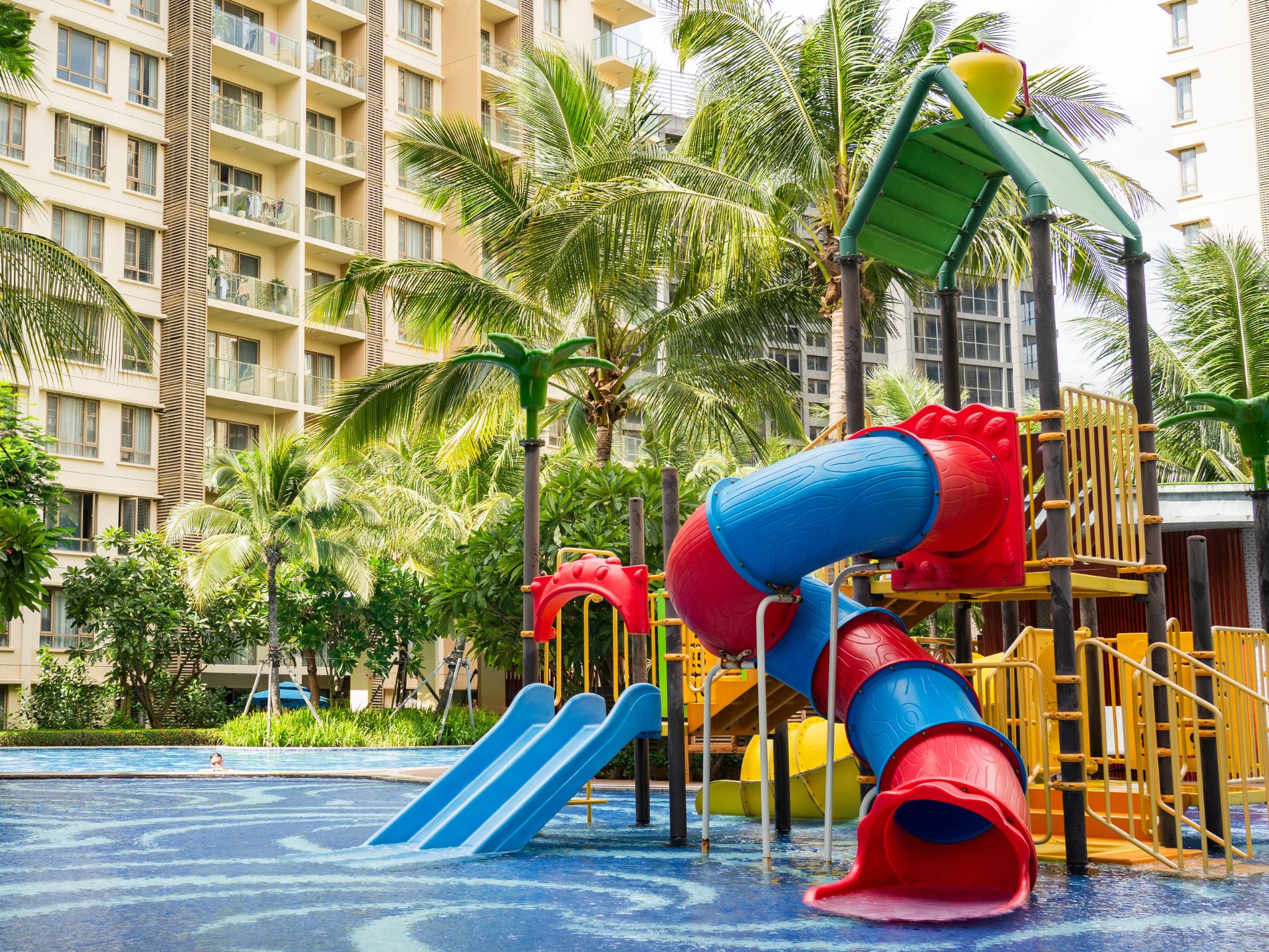 A water play area with slides and climbing structures, surrounded by palm trees and a hotel building.