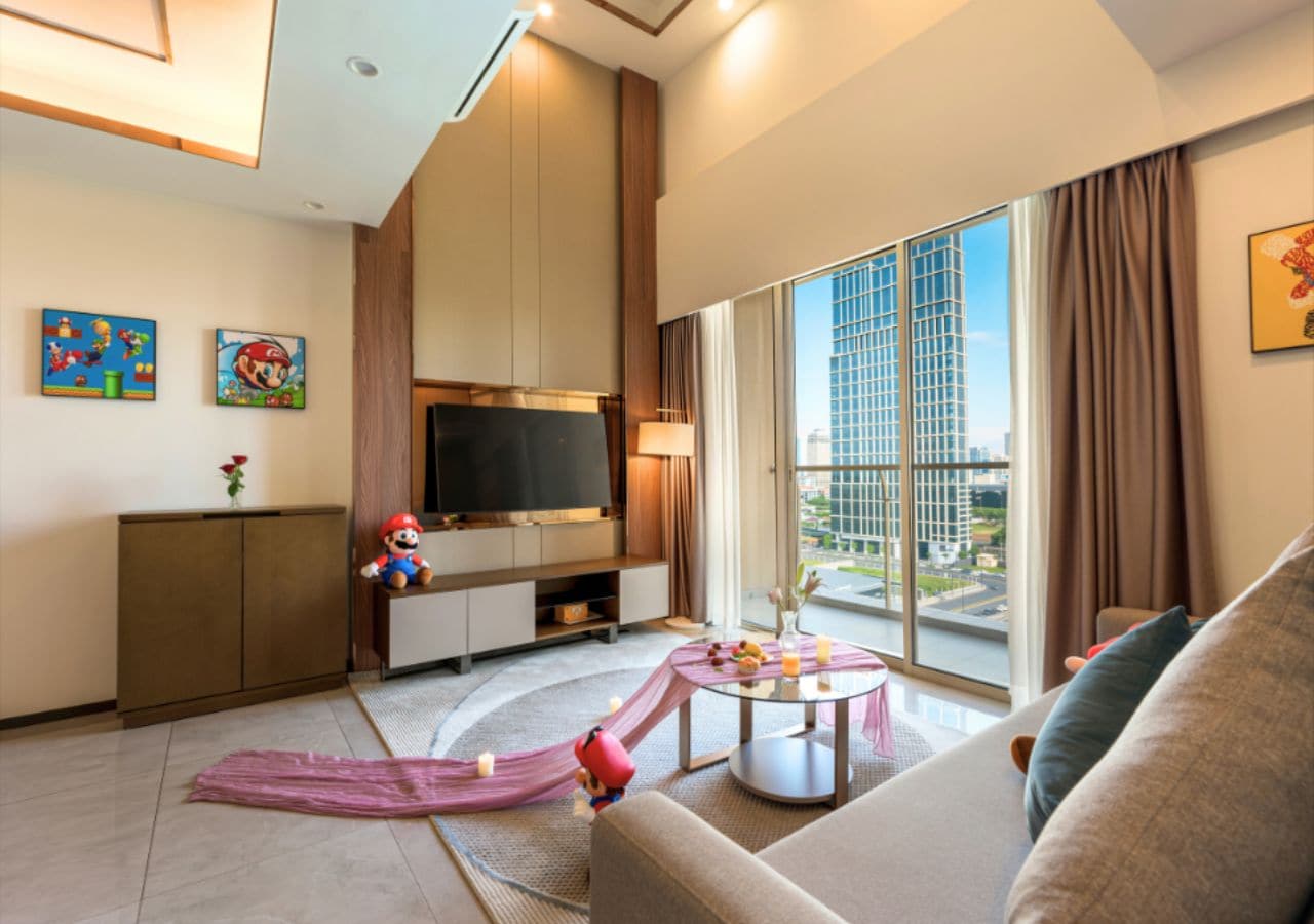 A modern hotel room with a large window, a flat-screen TV, a sofa, and a small round table with a pink tablecloth.