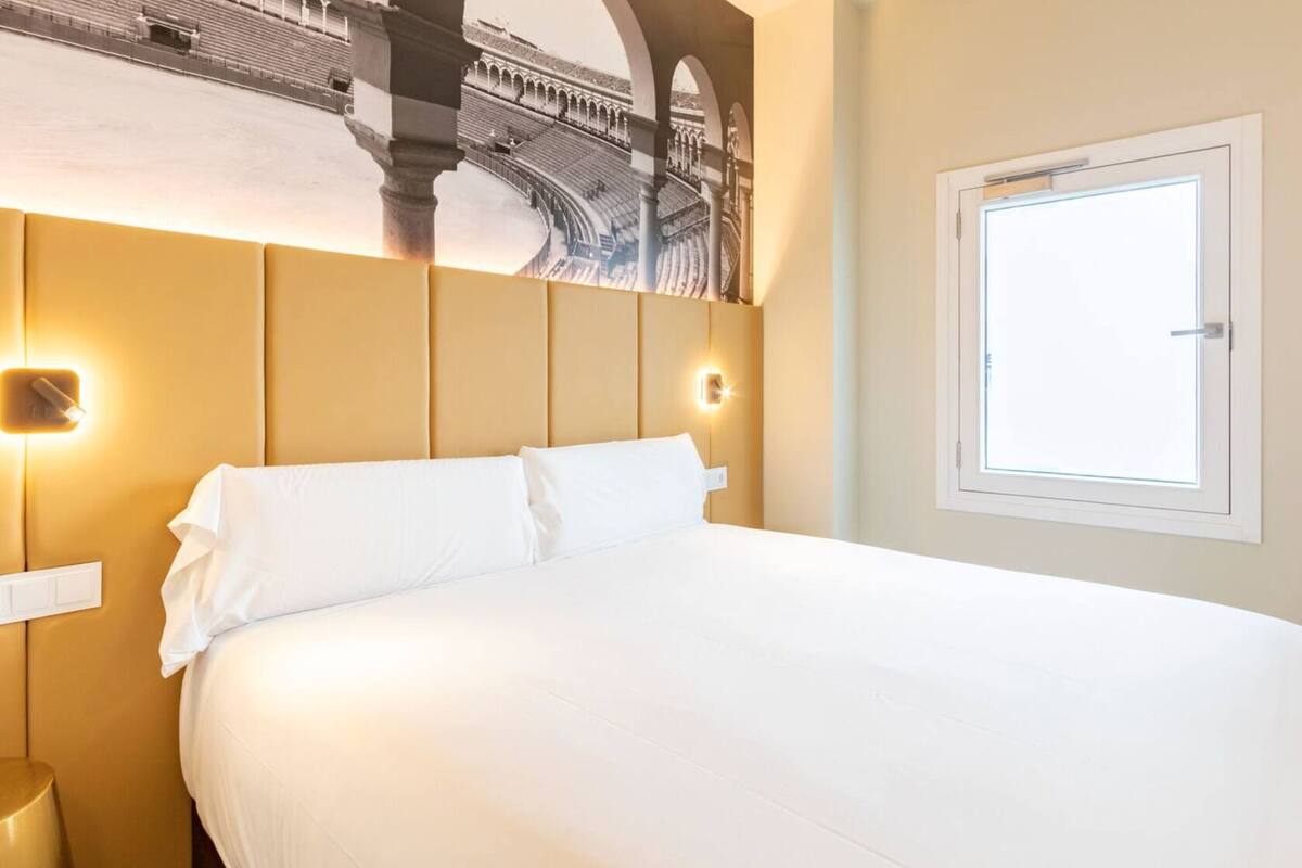 A hotel room with a large bed, a headboard with a mural, two wall-mounted lights, and a window.