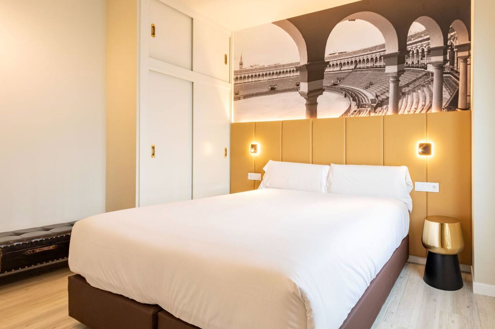 A hotel room with a large bed, a view of a historical building through an arched window, and a modern design with a wooden headboard.