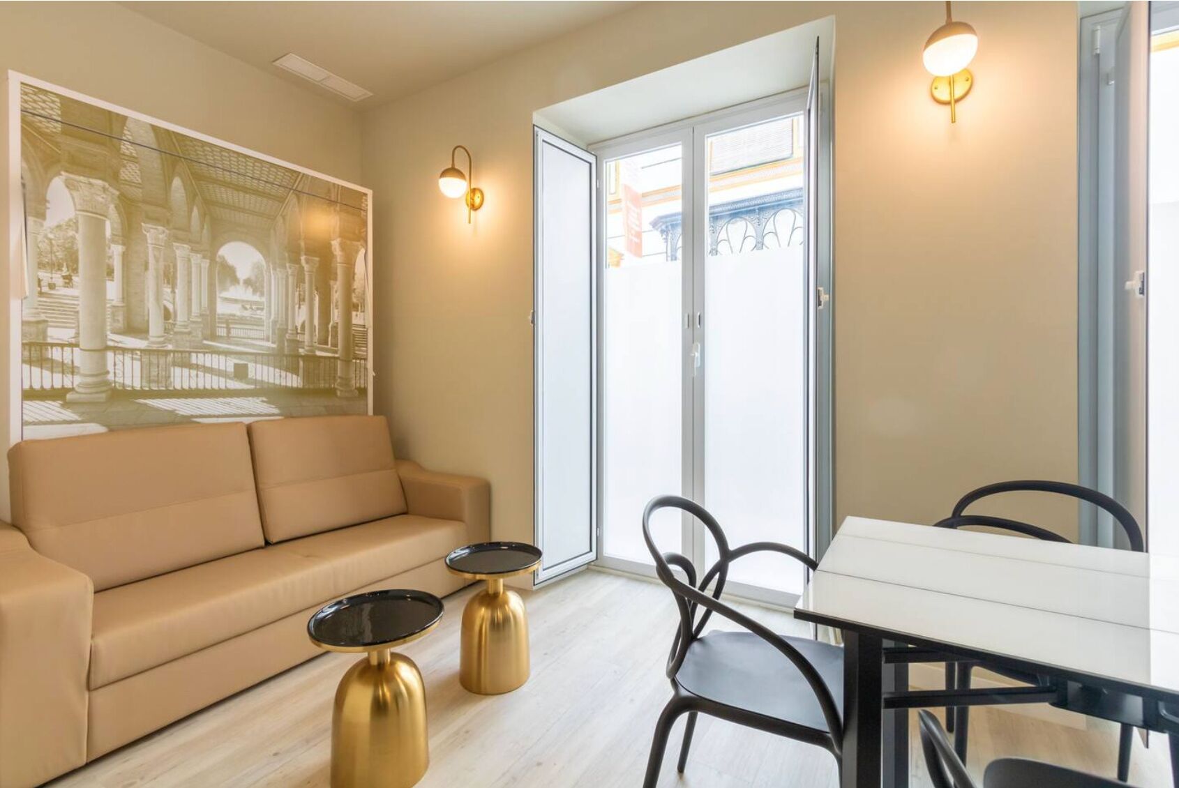 Una sala de estar con un sofá, dos mesitas doradas, una mesa de comedor con sillas y un ventanal con vistas a edificios.
