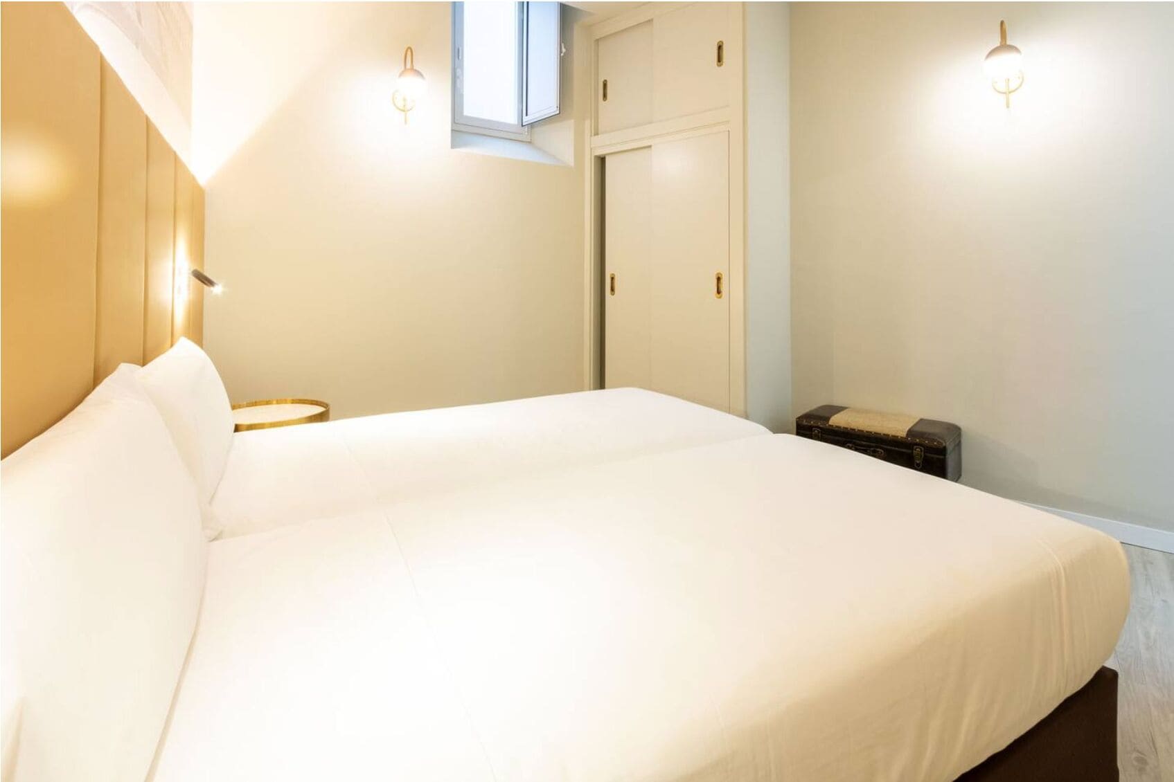 A neatly made bed in a room with a window, a wall-mounted light, and a small table with a suitcase.