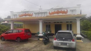 A building with a white facade and a large entrance. There are two vehicles parked in front: a red car and a silver car. - Hotel O Wisma Nadira Near SPBU PT. Trijaya Pugung PENENGAHAN (Pesisir Utara)