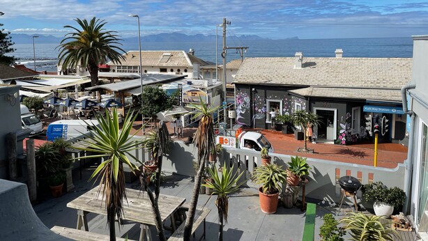 A coastal town with a view of the sea, featuring a street with shops, outdoor seating, and potted plants. - Studio apartment in the heart of Kalk Bay,  1 minute walk to the beach. (Cape Town)