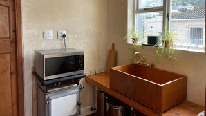 A kitchen with a microwave, a sink, and a window with a view of houses. - Studio apartment in the heart of Kalk Bay,  1 minute walk to the beach. (Cape Town)