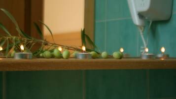 A bathroom with a green tiled wall, a wooden shelf with candles and green fruits, and a white electrical outlet.