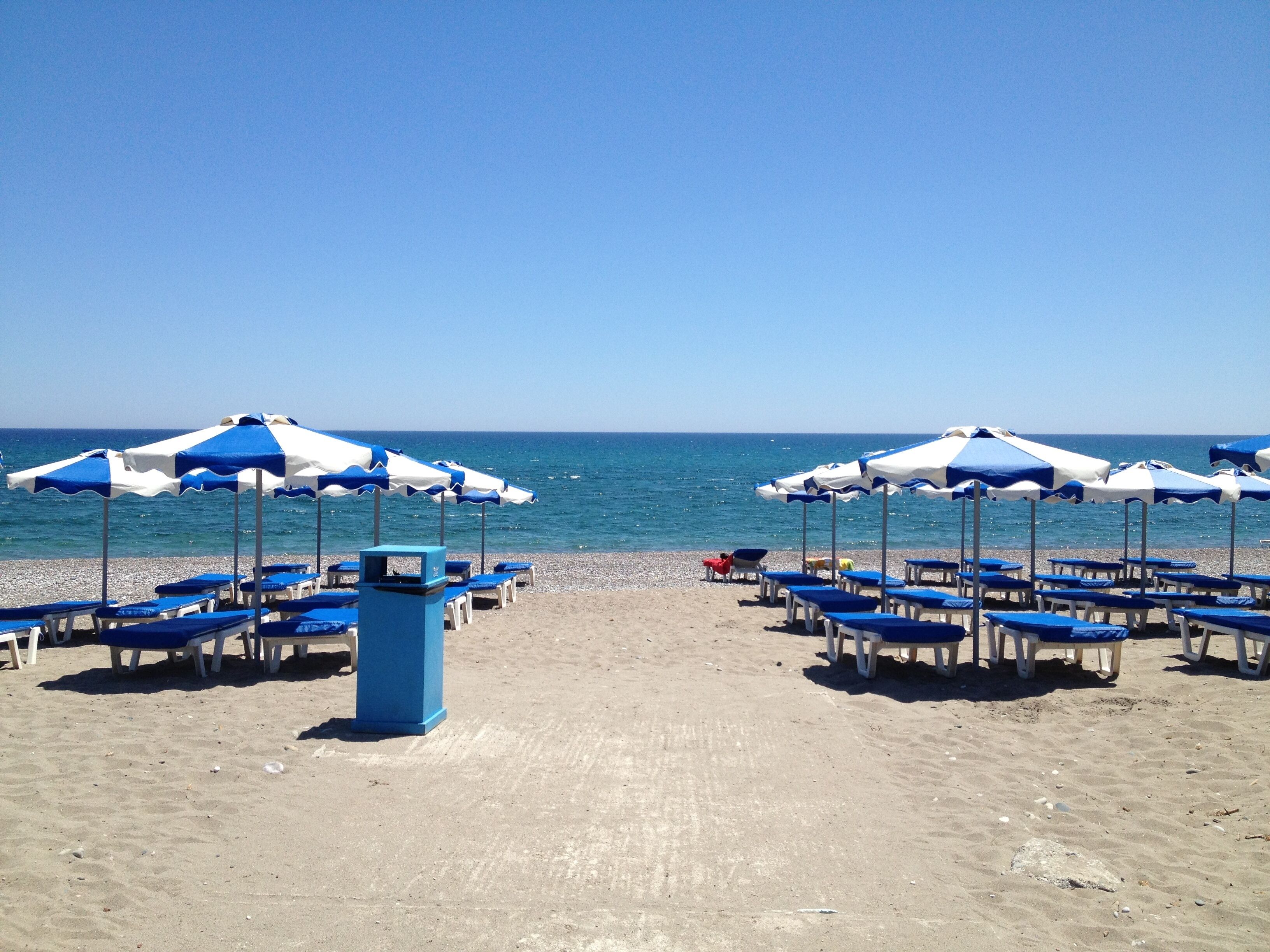 Una playa con hileras de sombrillas y sillas de playa azules y blancas orientadas al mar.