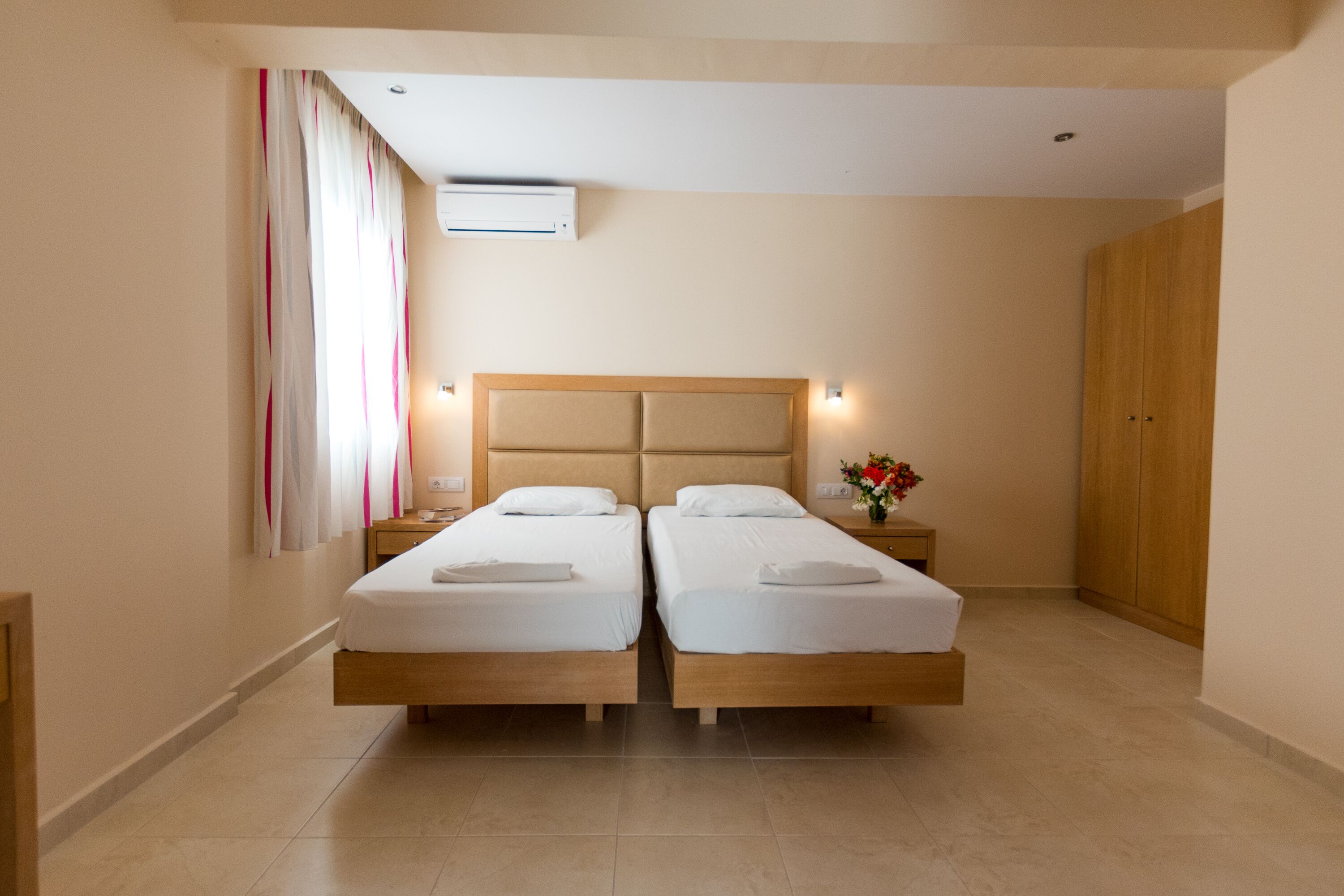 A twin room with two beds, a wooden wardrobe, a nightstand with a lamp, and a flower vase.