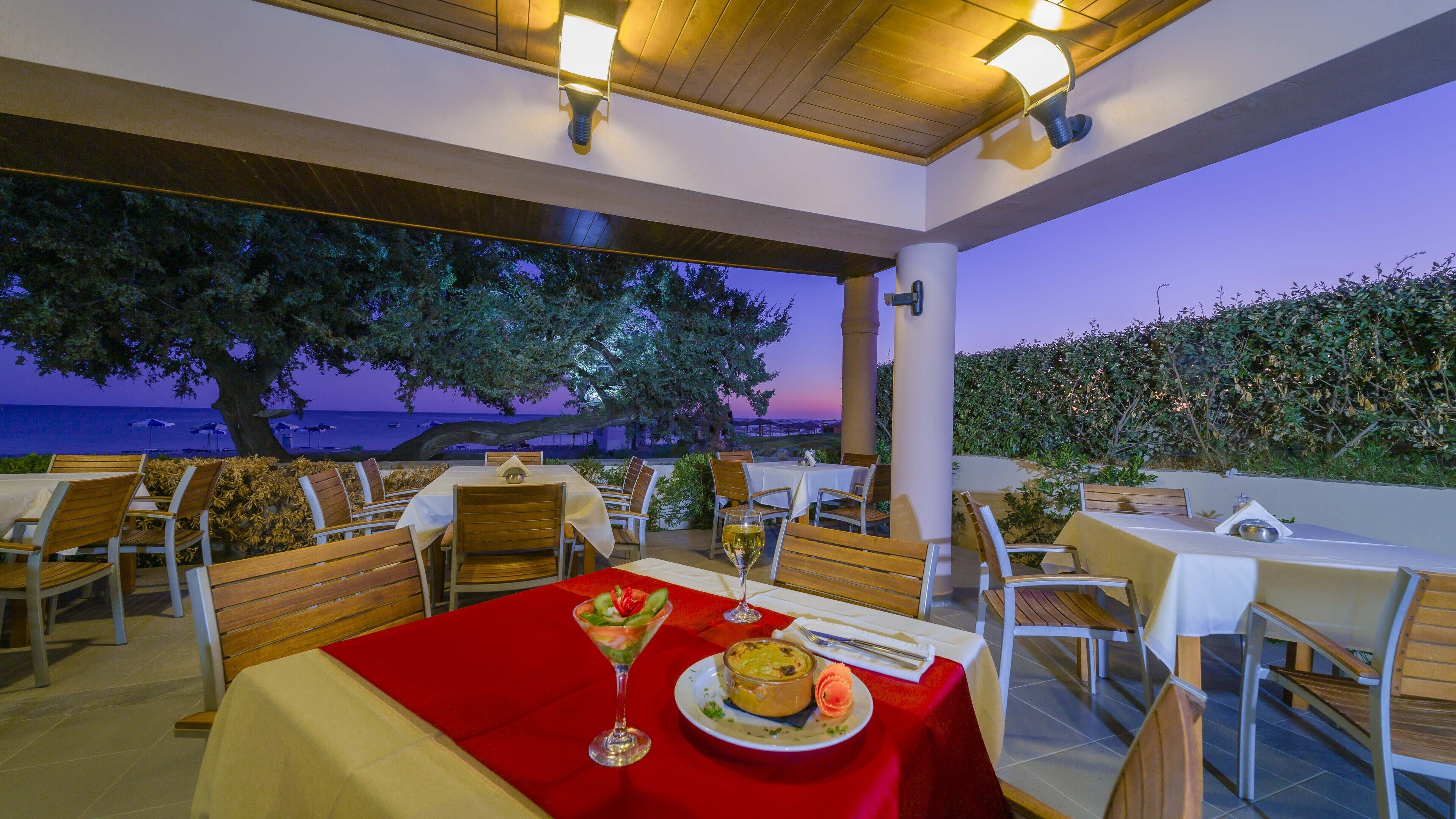 An outdoor dining area with tables and chairs, overlooking a body of water at dusk.