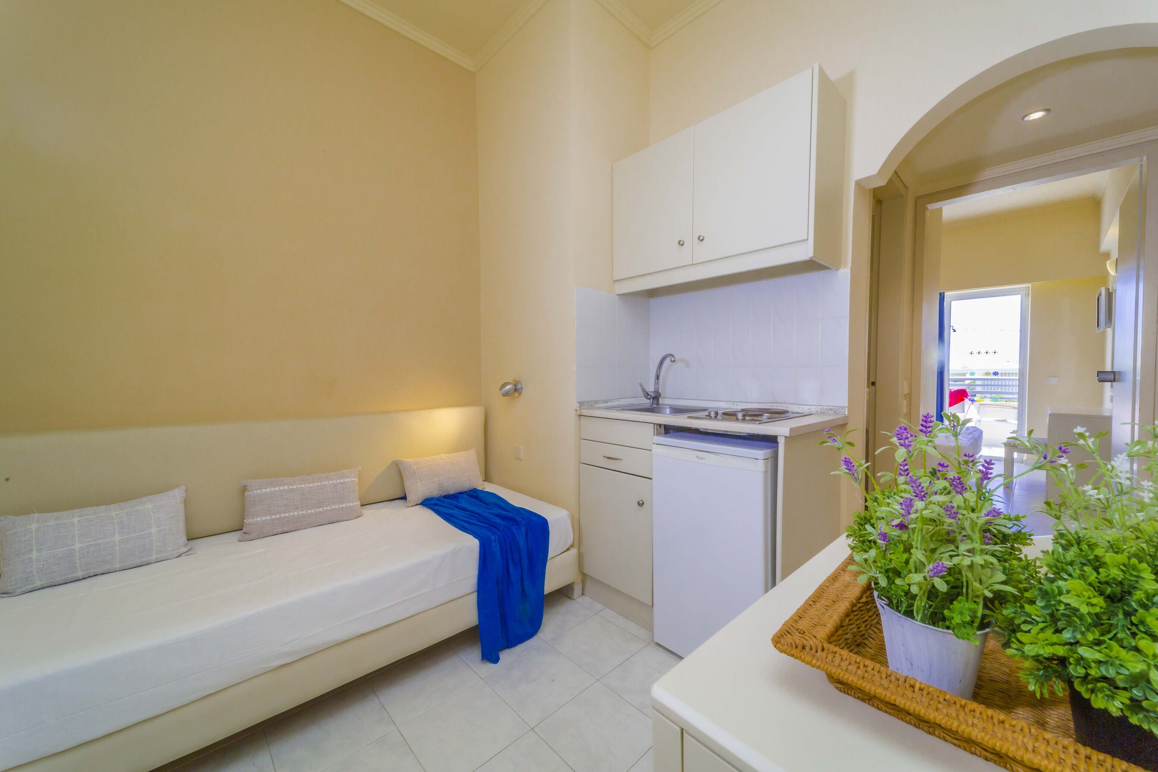 A compact room with a bed, a small kitchenette, and a potted plant.