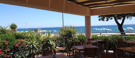 Eine Terrasse im Freien mit Holztischen und -stühlen, mit Blick auf einen Strand mit Sonnenschirmen und einem klaren, blauen Himmel.