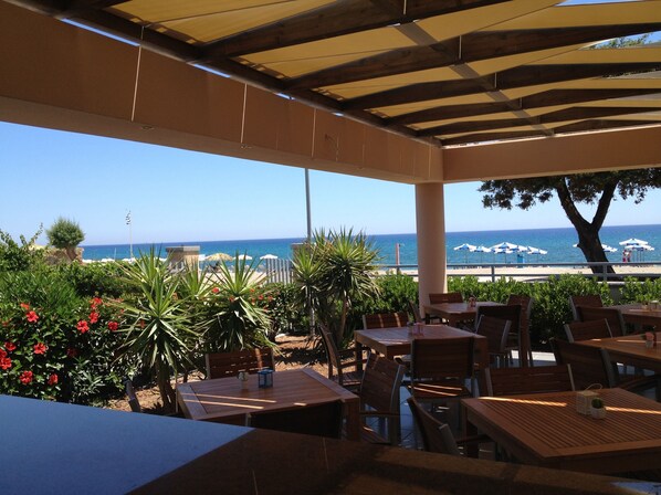 An outdoor patio with wooden tables and chairs, overlooking a beach with umbrellas and a clear blue sky.