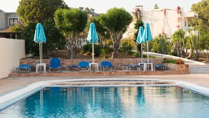 A swimming pool with blue lounge chairs and umbrellas, surrounded by trees and a stone wall. - Amaryllis Front Beach Hotel (Rhodes)