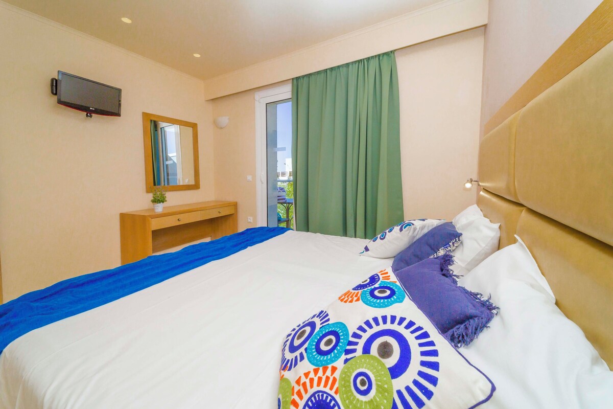 Habitación de hotel con cama, televisor montado en la pared, una pequeña mesa de madera y ventana con cortinas.