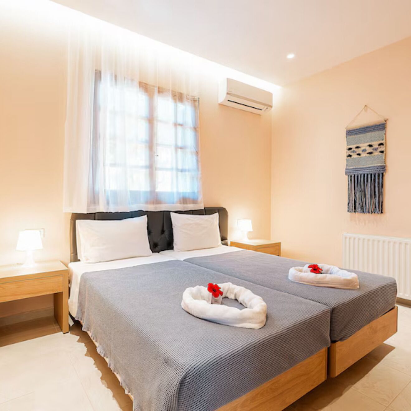 A double bed with a grey and white duvet, two bedside tables with lamps, a window with sheer curtains, an air conditioning unit, and a wall-mounted tapestry.