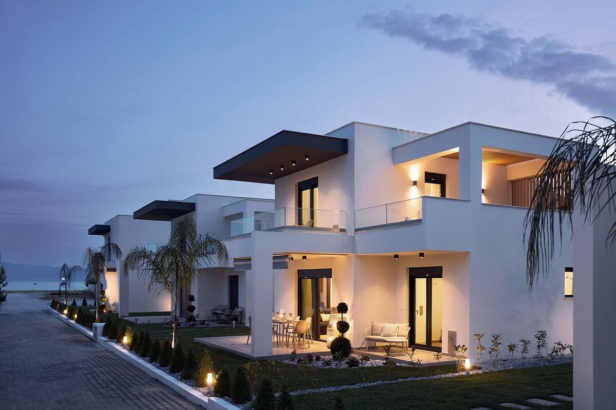 A modern two-story house with a balcony, outdoor seating, and a well-lit garden at dusk.