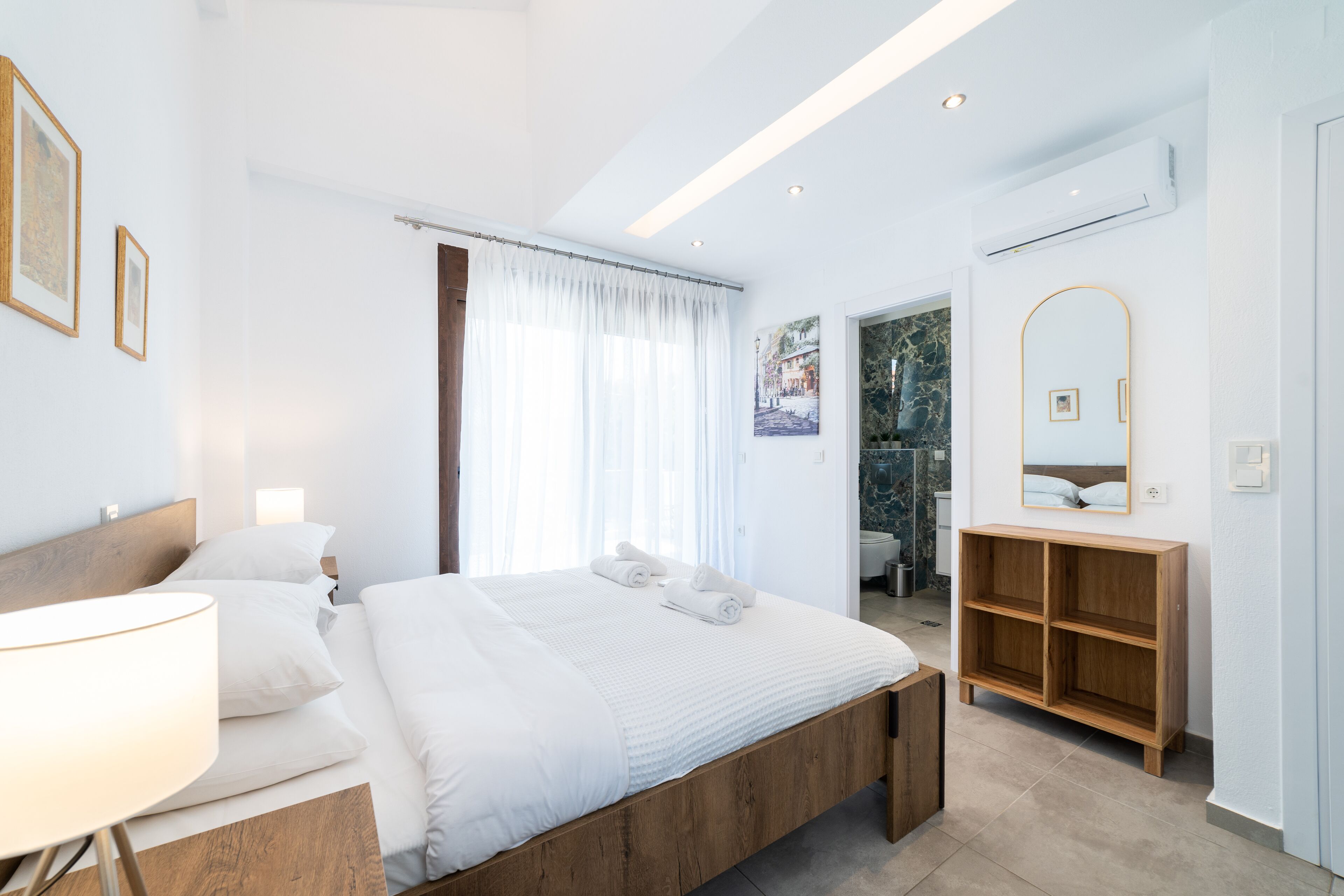 A modern hotel room with a wooden bed, a bedside lamp, a wardrobe, and a bathroom visible through an open door.
