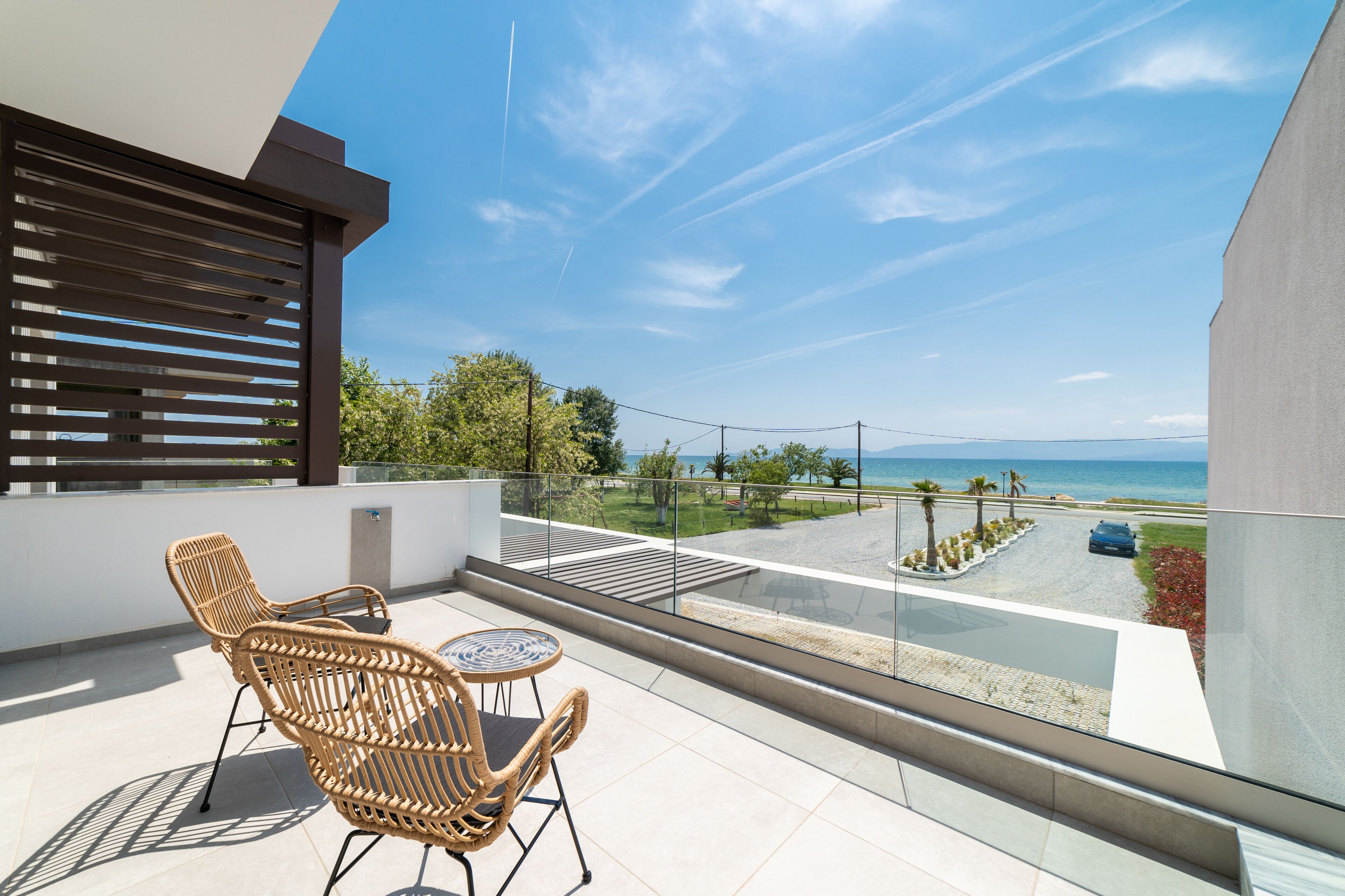 A balcony with wicker chairs and a small table, overlooking a paved area with a car and palm trees, under a clear blue sky.