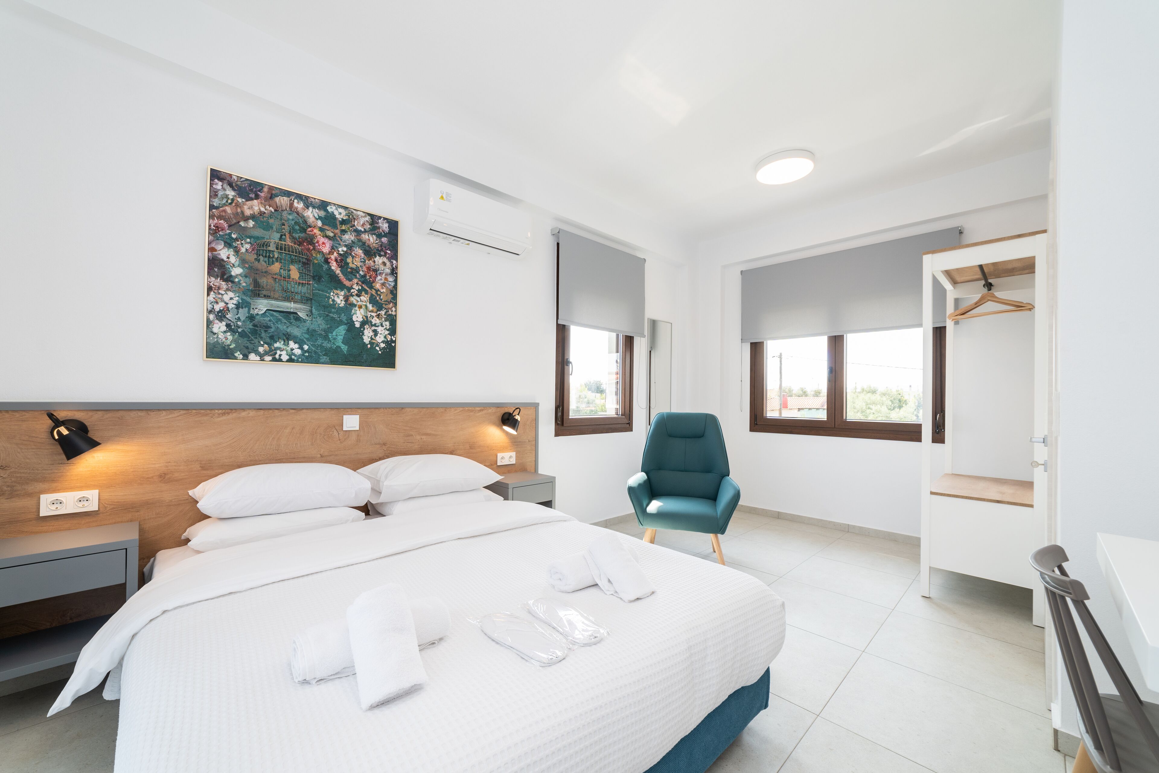 A modern hotel room with a large bed, a blue armchair, a painting on the wall, and a wardrobe with a mirror.