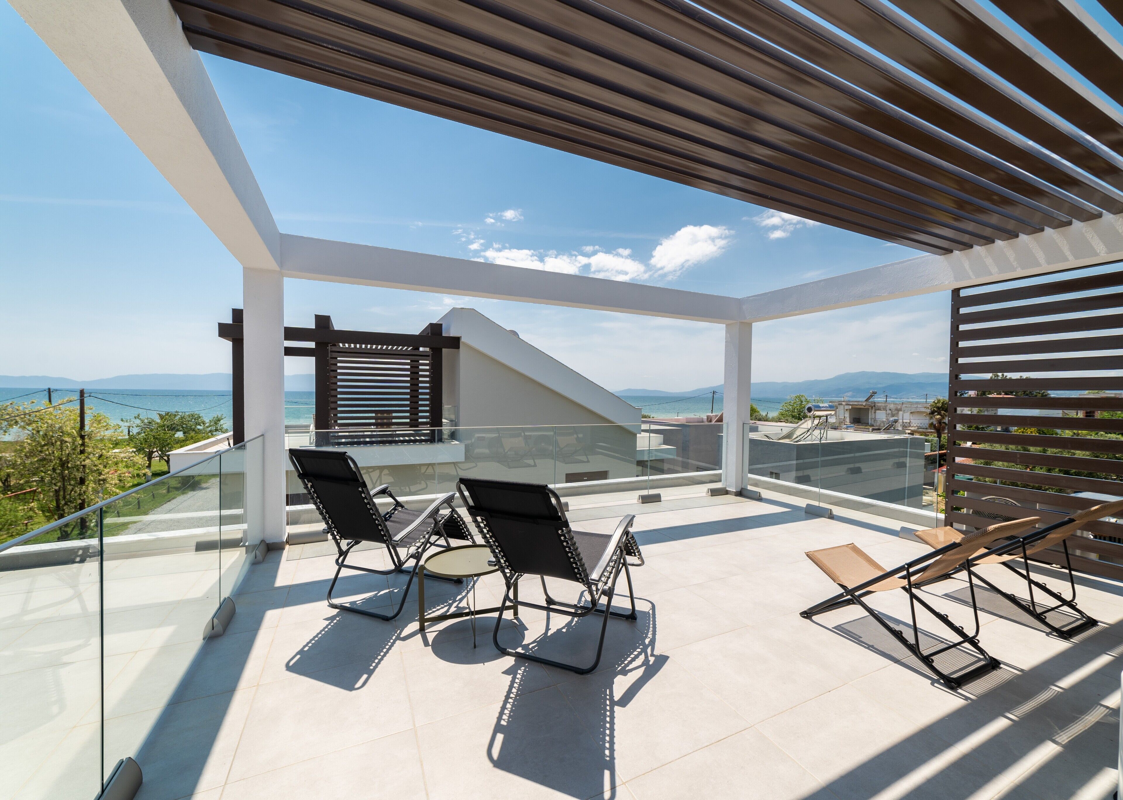 A modern outdoor balcony with glass railings, two black lounge chairs, and a wooden table.