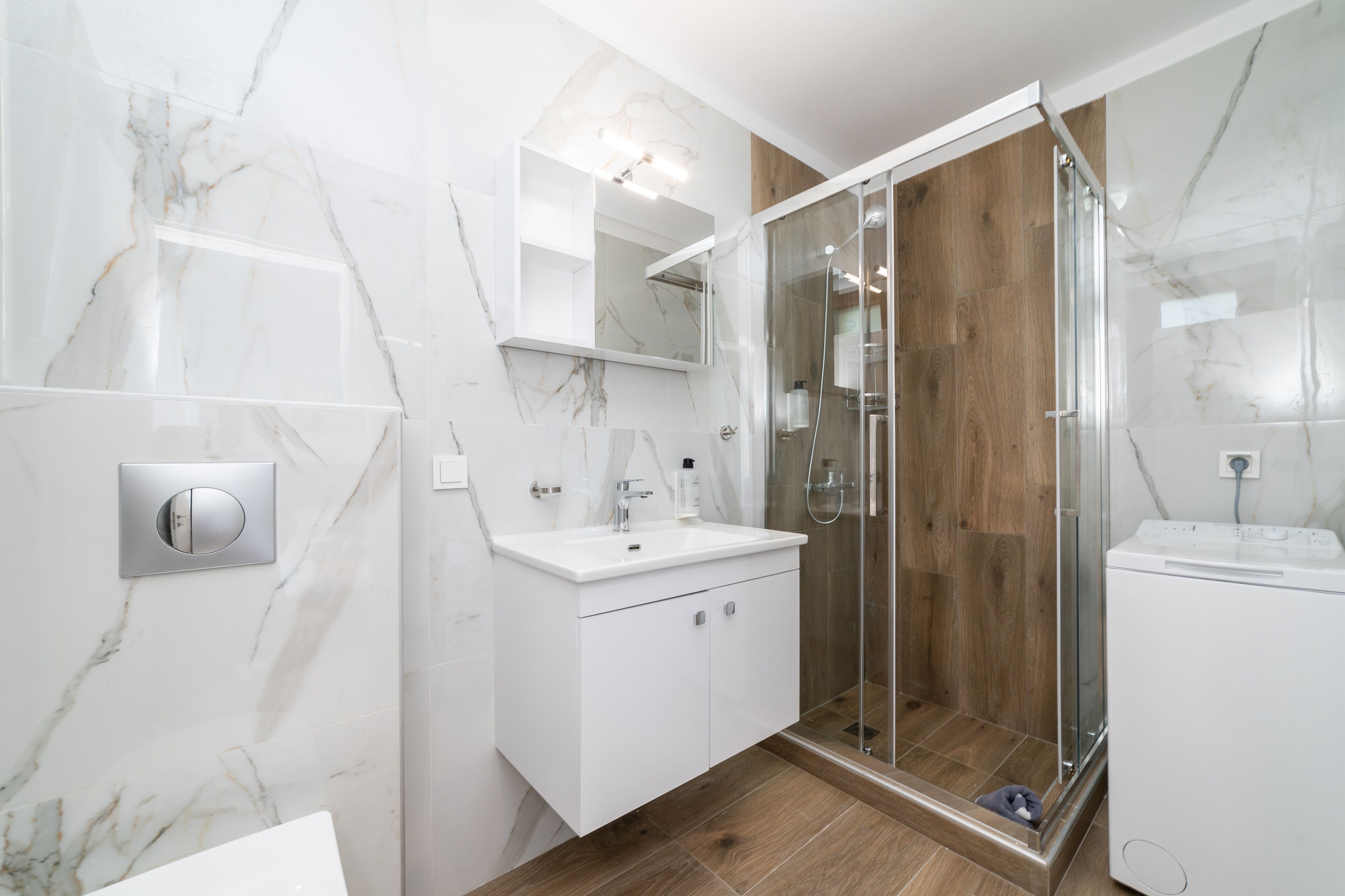 A modern bathroom with a marble wall, a wooden shower enclosure, and a built-in washing machine.