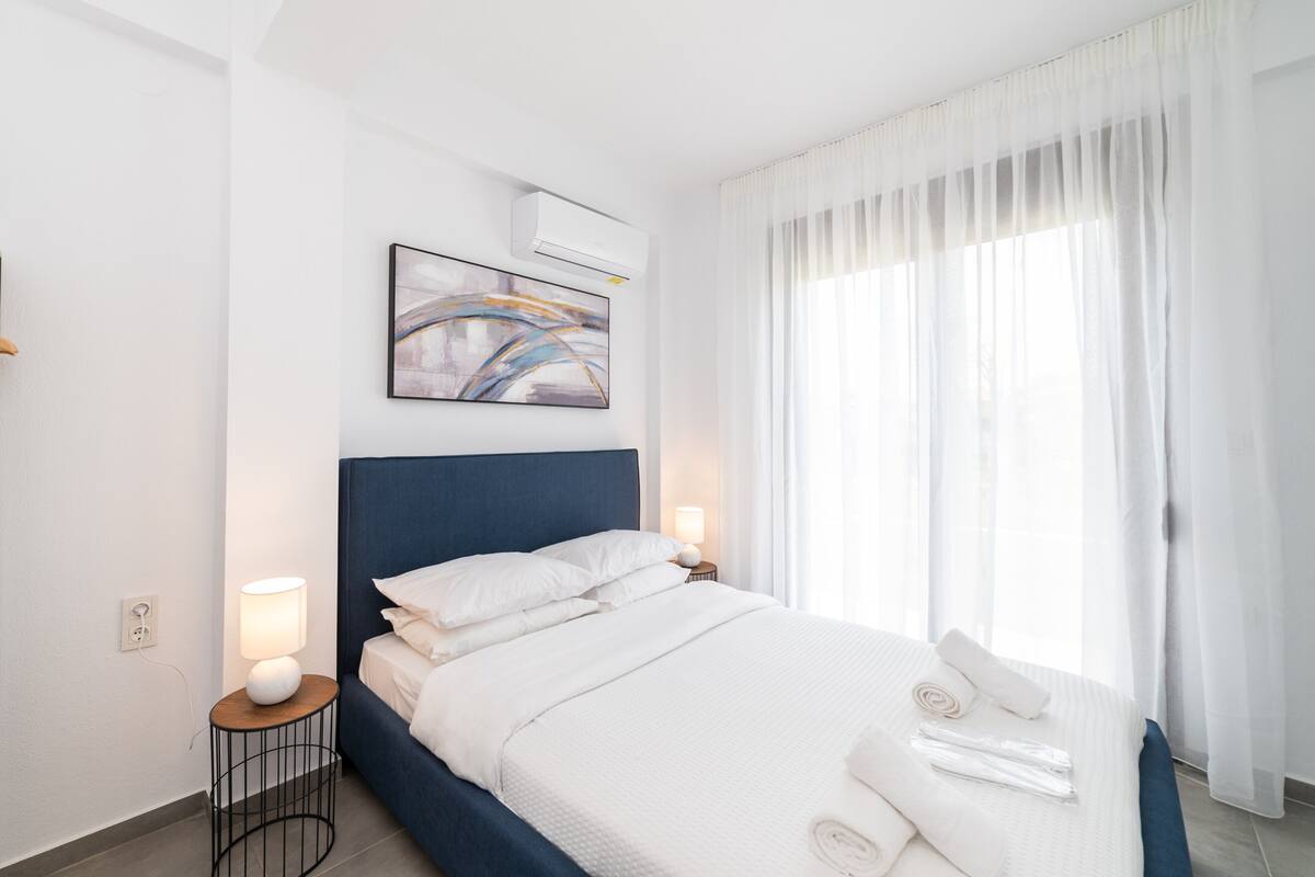 A modern bedroom with a large bed, white bedding, a blue headboard, a bedside table with a lamp, and a window with sheer curtains.