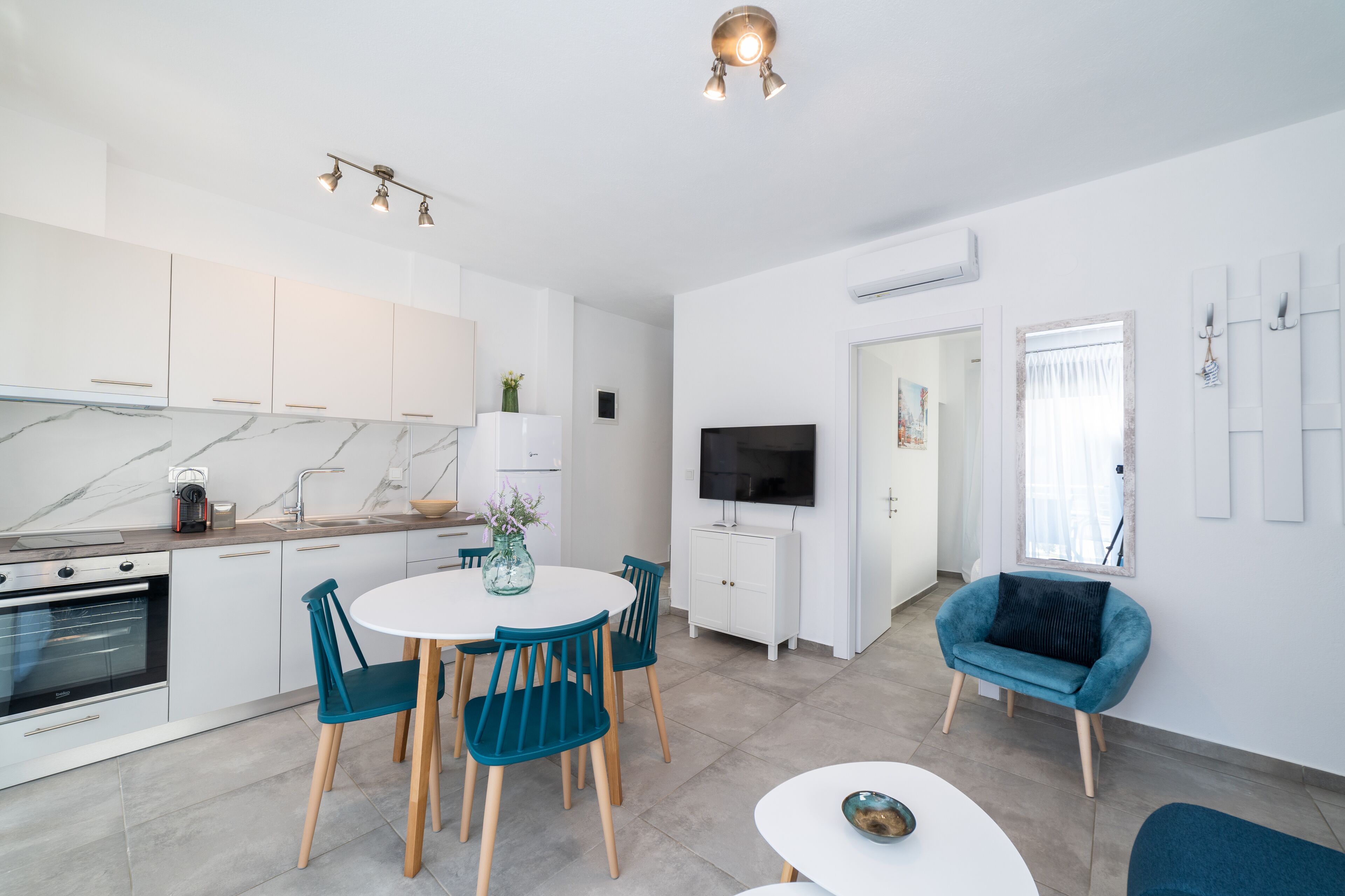A modern kitchen with white cabinets, a marble countertop, and a built-in oven. There is a dining area with a white table and teal chairs. A flat-screen TV is mounted on the wall, and a blue armchair is visible in the background.