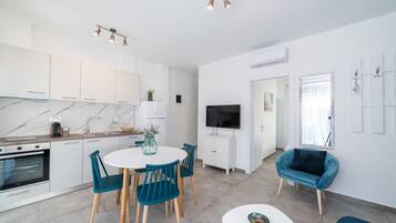 A modern kitchen with white cabinets, a marble countertop, and a built-in oven. There is a dining area with a white table and teal chairs. A flat-screen TV is mounted on the wall, and a blue armchair is visible in the background.